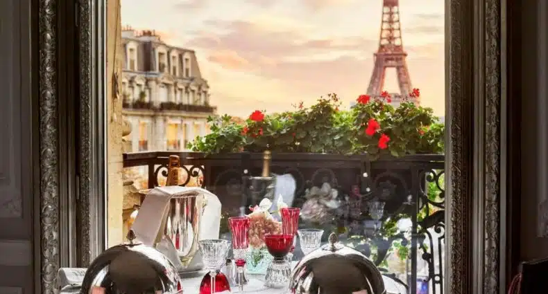 Uma mesa para dois com pratos cobertos e copos vermelhos tem vista para a Torre Eiffel ao pôr do sol por uma janela aberta, no Hôtel Plaza Athénée - Dorchester Collection, para representar hotéis românticos em Paris