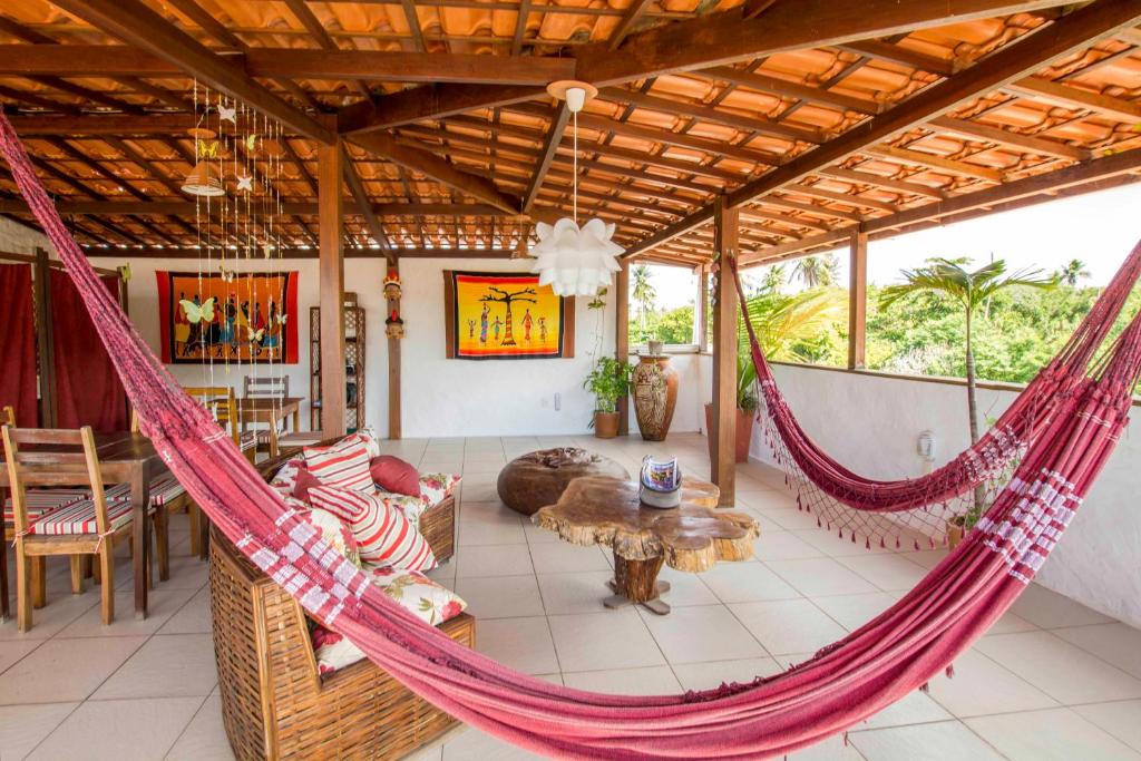 Pátio coberto com telhado de terracota, redes vermelhas, móveis de madeira, arte na parede, plantas e uma mesa de madeira rústica, na propriedade da Pousada Borboleta.