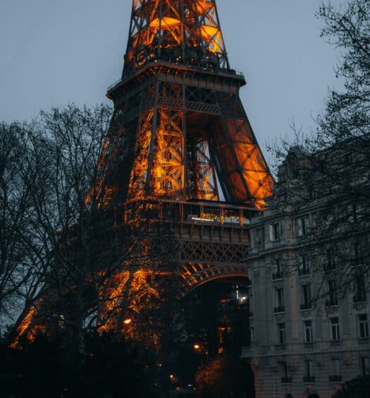 A base da Torre Eiffel iluminada ao anoitecer, com árvores nuas e um prédio próximo em primeiro plano. Ilustra o post de eSIM Paris.