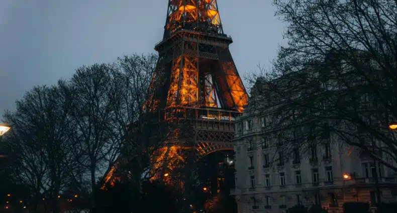 A base da Torre Eiffel iluminada ao anoitecer, com árvores nuas e um prédio próximo em primeiro plano. Ilustra o post de eSIM Paris.