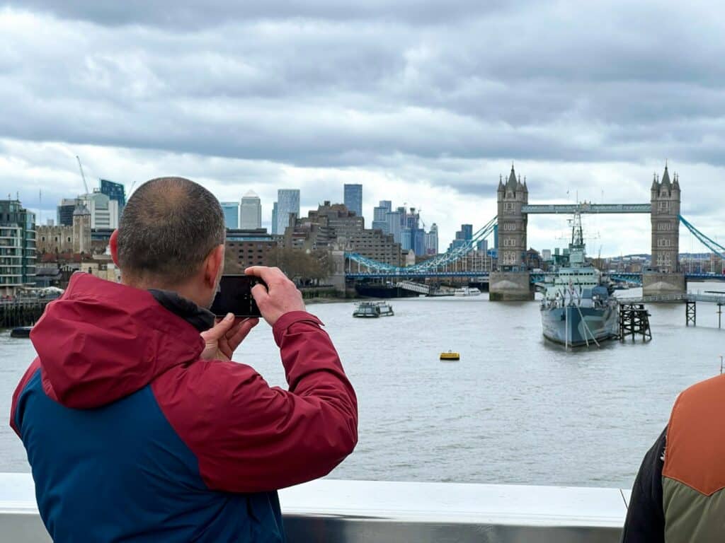 Uma pessoa com uma jaqueta vermelha e azul tira uma foto da Tower Bridge e do rio Tâmisa em um dia nublado em Londres. Ilustra o post de eSIM Inglaterra.