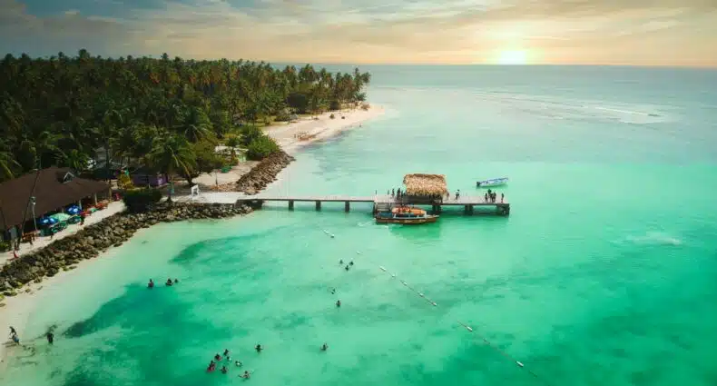 Vista aérea de Pigeon Point Beach, praia tropical em Trindade e Tobago com um píer, barcos, pessoas nadando e palmeiras ao pôr do sol. A foto ilustra o post de eSIM em Trindade e Tobago.