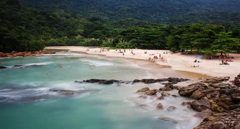 Pessoas relaxam na Praia do Meio em Trindade, com rochas em primeiro plano e uma densa floresta verde e colinas ao fundo, para representar pousadas em Trindade.