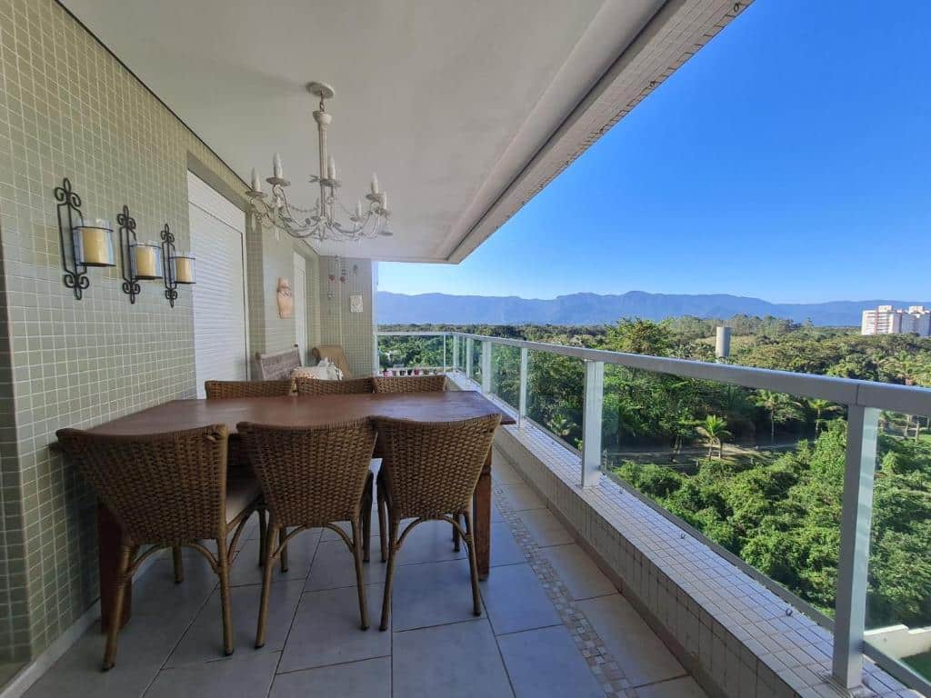 Uma varanda com cadeiras de vime ao redor de uma mesa de madeira tem vista para árvores e montanhas distantes sob um céu azul claro, na propriedade do Riviera São Lourenço Excelente Apartamento e Local.