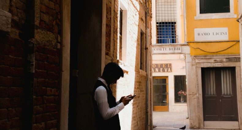 Uma pessoa está em um beco estreito e ensolarado, olhando para um telefone, com prédios e placas de hotéis ao fundo em Veneza, que pode ser visitada com o eSIM Itália.