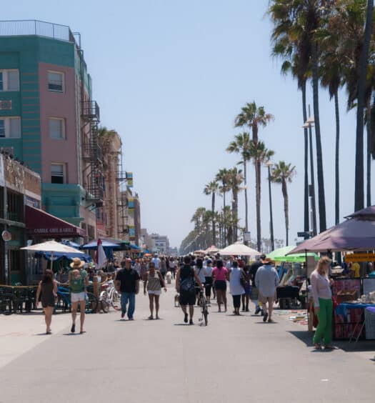 As pessoas caminham por um calçadão movimentado de Venice Beach, repleto de lojas, palmeiras e barracas de vendedores em um dia ensolarado. Imagem para ilustrar post sobre eSIM Los Angeles.