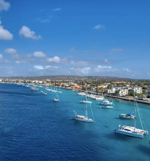 Vários veleiros e iates ancorados em águas azuis e cristalinas perto de uma cidade costeira sob um céu quase limpo com nuvens esparsas. Imagem para ilustrar post sobre eSIM Bonaire.