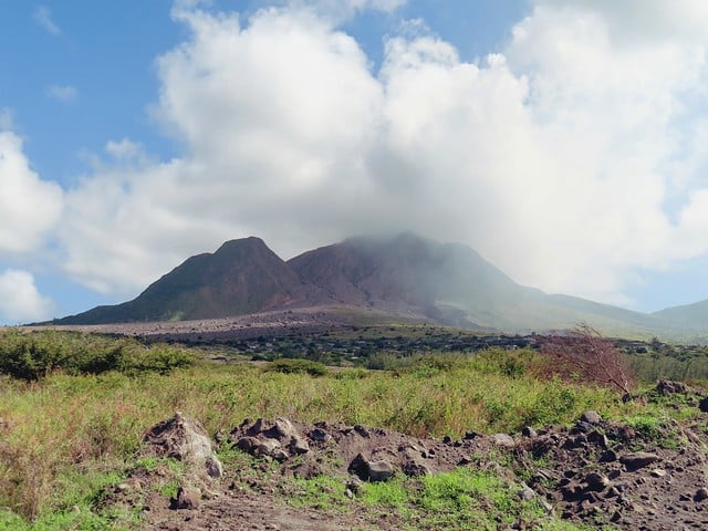 Vulcão Soufrière Hills, em Montserrat, com um pico coberto de nuvens, cercado por vegetação verde e terreno rochoso sob um céu parcialmente nublado. A foto faz parte do post de eSIM Montserrat.