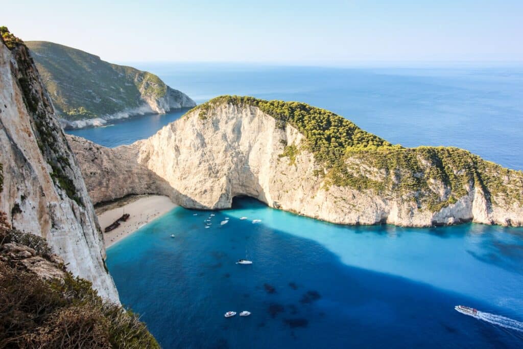 Vista aérea de uma enseada com praia de areia, águas azul-turquesa, barcos ancorados e penhascos rochosos com vegetação em Zakynthos, que pode ser visitada com o eSIM Grécia.