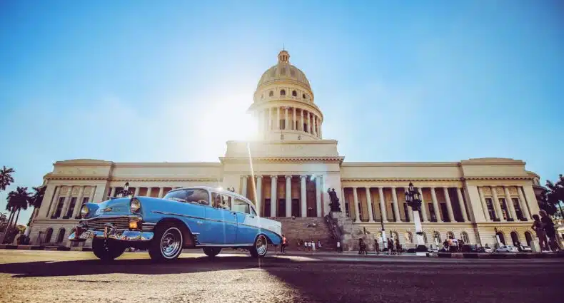 Um carro azul clássico está estacionado em frente a um grande prédio governamental abobadado o Capitólio de Havana, sob um céu claro e brilhante que pode ser visitado com o eSIM Cuba.