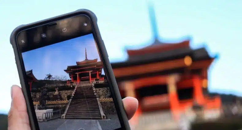 Uma mão segura um telefone tirando uma foto do Fushimi Inari-taisha em Kyoto Japão, um templo vermelho com escadas, com um templo desfocado visível ao fundo, que pode ser visitado com o eSIM Ásia.
