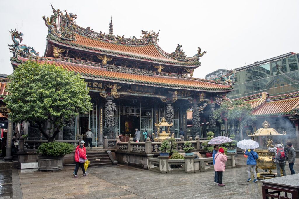 Pessoas com guarda-chuvas em Taipei, Taiwan caminham em frente a um templo tradicional ornamentado em um dia chuvoso, com fumaça de incenso em primeiro plano, que pode ser visitado com um eSIM Ásia.