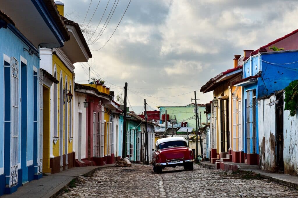 Um carro clássico vermelho passa por uma rua de paralelepípedos ladeada por casas coloridas sob um céu nublado em Trinidad que pode ser visitado com o eSIM Cuba.