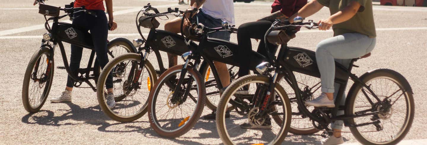 Três pessoas estão andando de bicicletas elétricas pretas em um dia ensolarado, com as pernas visíveis e as mãos no guidão. Imagens para ilustrar post sobre aluguel de bicicleta elétrica em Lisboa.