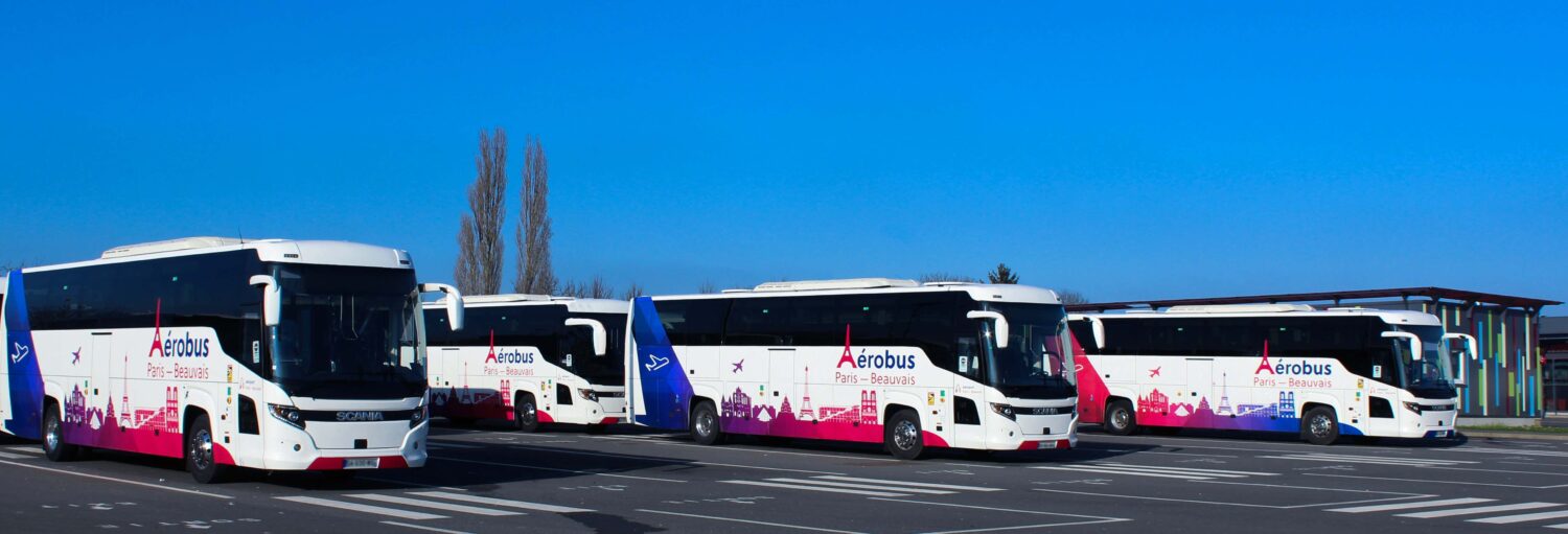 Quatro ônibus Aerobus brancos e rosas estão estacionados em fila em um estacionamento pavimentado sob um céu azul claro. A imagem ilustra o post sobre ônibus entre o aeroporto de Beauvais e Paris.