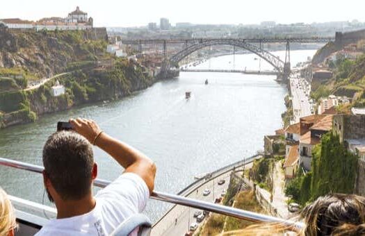 Pessoas em um passeio de ônibus de dois andares com vista para um rio, uma ponte e uma paisagem urbana em um dia ensolarado. A foto é capa do post de Ônibus turístico do Porto, passeio de barco e adega Burmester.