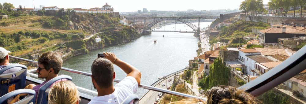 Pessoas em um passeio de ônibus de dois andares com vista para um rio, uma ponte e uma paisagem urbana em um dia ensolarado. A foto é capa do post de Ônibus turístico do Porto, passeio de barco e adega Burmester.