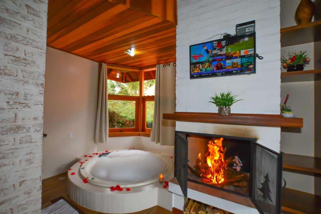 Uma banheira de hidromassagem redonda perto de uma lareira acesa, com pétalas de rosa na borda da banheira e uma TV montada acima da lareira, em um dos quartos da Pousada Cantinho de Monte Verde.