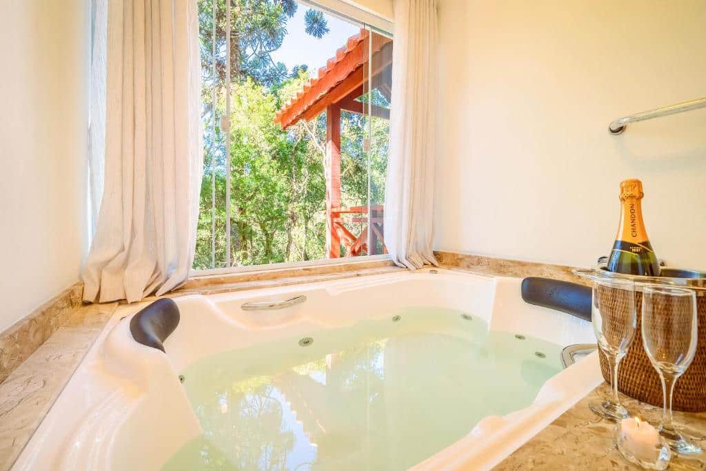 Uma banheira de hidromassagem cheia ao lado de uma janela com vista para a floresta, uma garrafa de champanhe em um balde de gelo e duas taças na borda, em um dos quartos da VELINN Pousada Jardim da Mantiqueira.