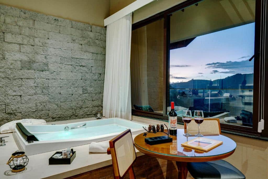 Jacuzzi interna ao lado de uma mesa redonda com vinho e duas taças, janela grande com vista para o pôr do sol em colinas distantes, em um dos quartos da Pousada Spa Mirante da Colyna.
