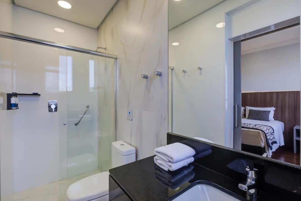 Banheiro moderno com chuveiro de vidro, vaso sanitário, bancada preta, toalhas empilhadas e vista de um quarto através de uma porta aberta, em um dos quartos do Slaviero Essential Blumenau.