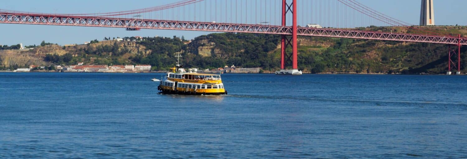 Uma balsa amarela atravessa um rio azul com uma ponte suspensa vermelha e colinas verdes ao fundo. Imagem para ilustrar post sobre barco turístico de Lisboa.