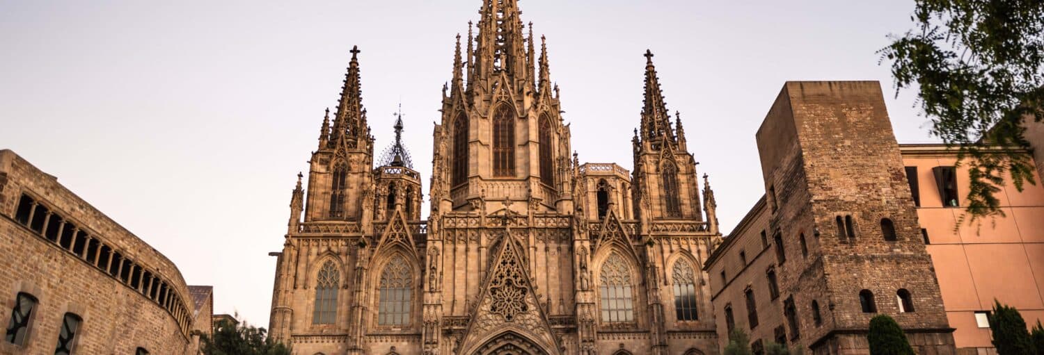 Catedral em estilo gótico, com torres altas e trabalhos intrincados em pedra, cercada por edifícios mais antigos ao anoitecer. A imagem ilustra o post sobre Tour pelos bairros de Barcelona.