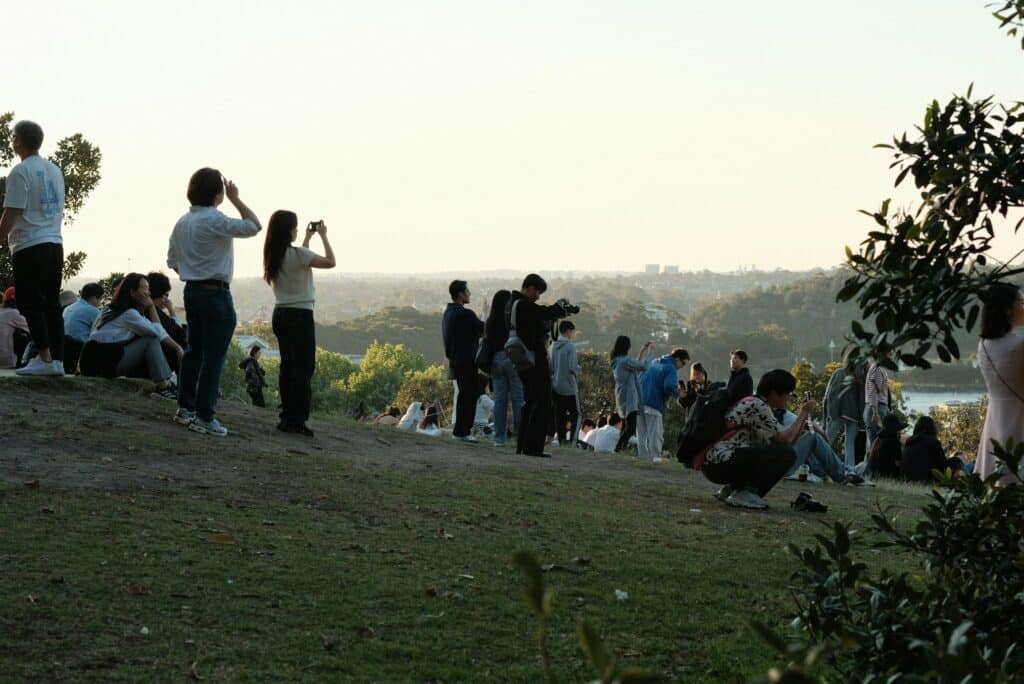 Um grupo de pessoas fica em pé, senta e tira fotos em uma colina gramada do Observatory Hill, em Sydney, com vista para árvores e um corpo d'água ao longe. A foto faz parte do post de eSIM Sydney.