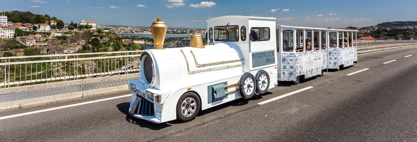 Um trem turístico branco com detalhes em dourado passa por uma ponte com uma paisagem urbana ao fundo sob um céu claro. A foto é capa do post Visita à adega Real Companhia Velha + Trem turístico.