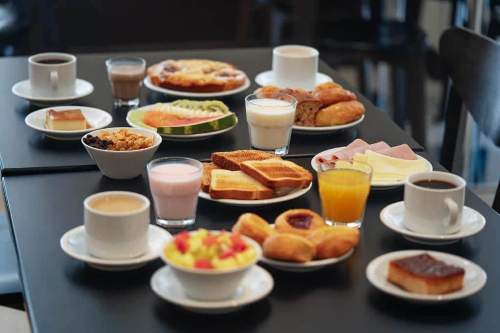 Uma mesa posta com café, suco, leite, torradas, doces, frutas, cereais e frios em pratos e copos brancos, servido no Mérit San Telmo.