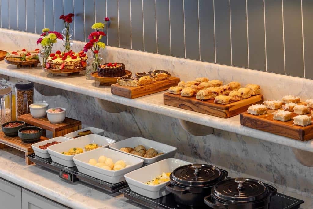 Um bufê com vários doces, sobremesas e pratos quentes dispostos em uma bancada de mármore e bandejas de madeira, servido no Villano Hotel.