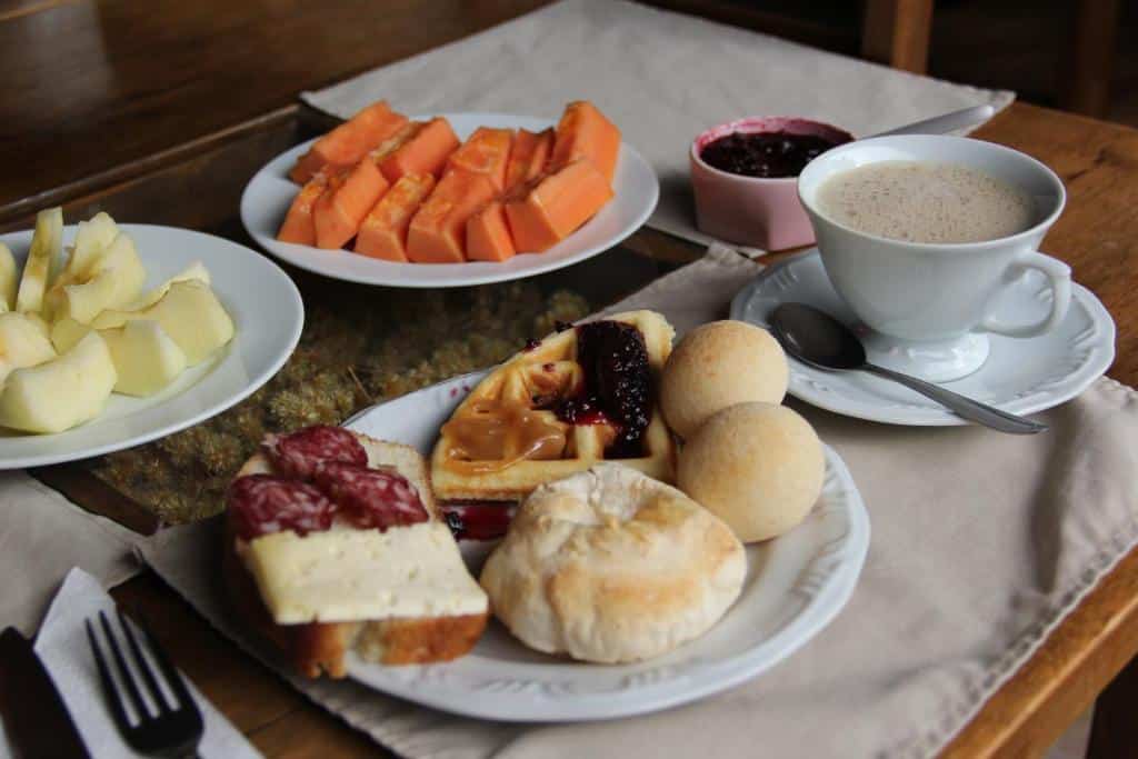 Um café da manhã com frutas, pães, queijos, carnes, bolos, geleia e uma xícara de café em um jogo de mesa, servido no Café e Cabans Lenha no Fogo.