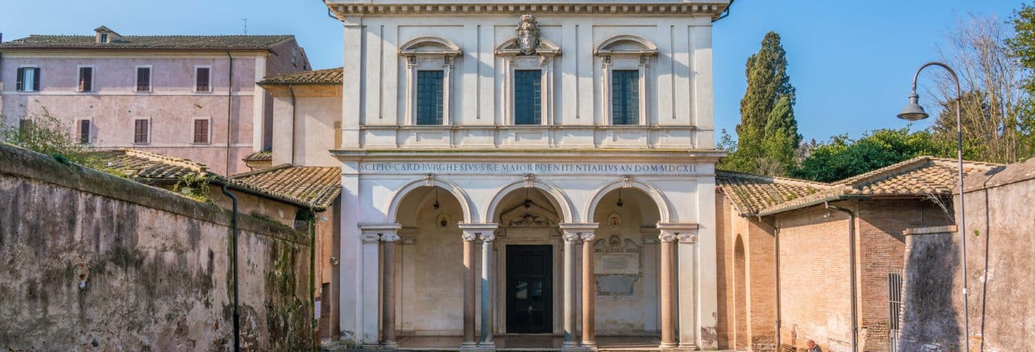 Uma igreja histórica de pedra branca com três entradas em arco e colunas, ladeada por antigos edifícios de tijolos e árvores no Tour pelas catacumbas de San Sebastiano + Ônibus saindo de Roma Termini.