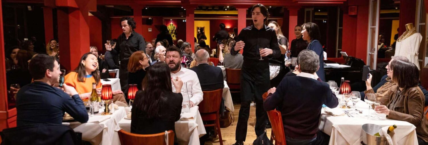 Restaurante lotado com clientes em mesas, um homem de pé cantando, e pessoas comendo, bebendo e conversando, ilustrando o passeio de jantar com espetáculo de ópera no Bel Canto