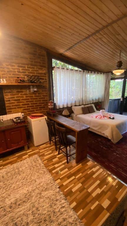 Um quarto estúdio aconchegante com uma cama, uma pequena área de cozinha, frigobar, teto de madeira e janelas grandes com cortinas transparentes.