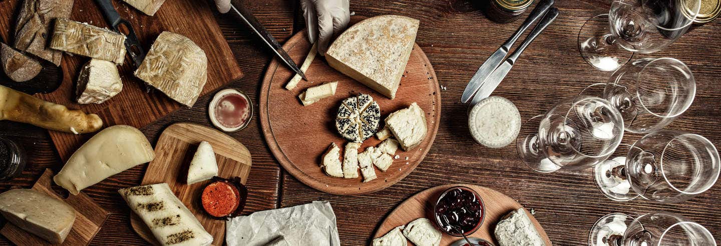 Queijos variados, pães, pastas e taças de vinho vazias dispostas sobre uma mesa de madeira, numa perspectiva de cima para baixo, ilustrando a capa do passeio de degustação de vinhos e queijos