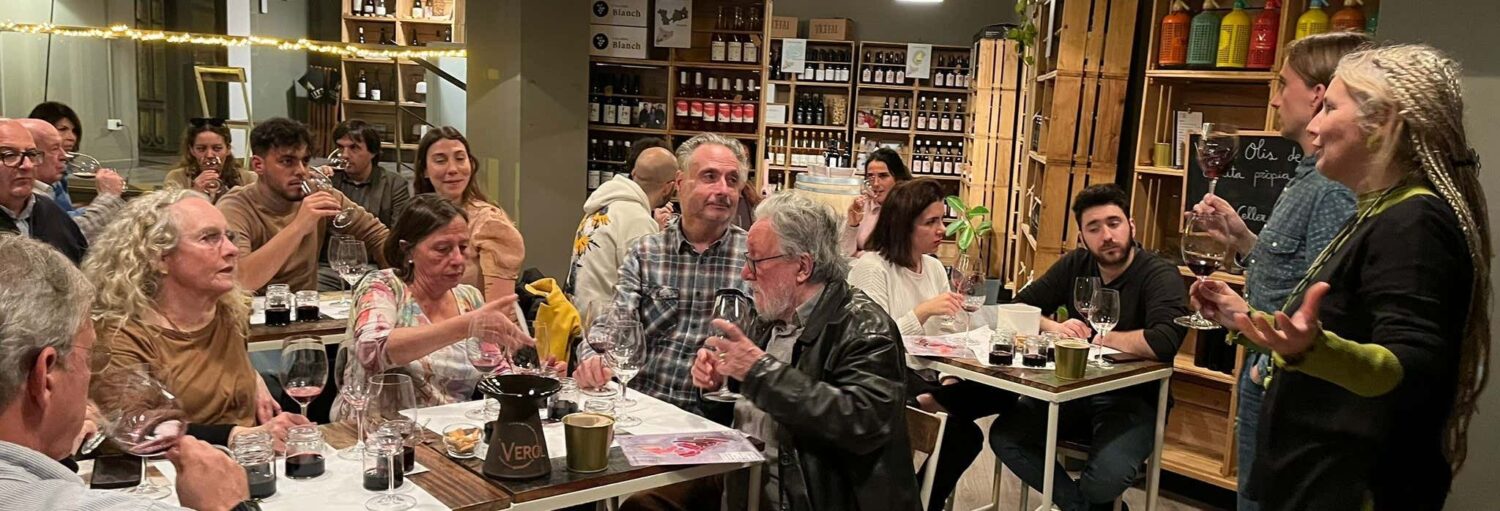 Pessoas sentadas em mesas em uma loja de vinhos, degustando vinhos e ouvindo duas pessoas de pé e falando na frente. A imagem ilustra o post sobre degustação de vinhos e queijos em Barcelona.