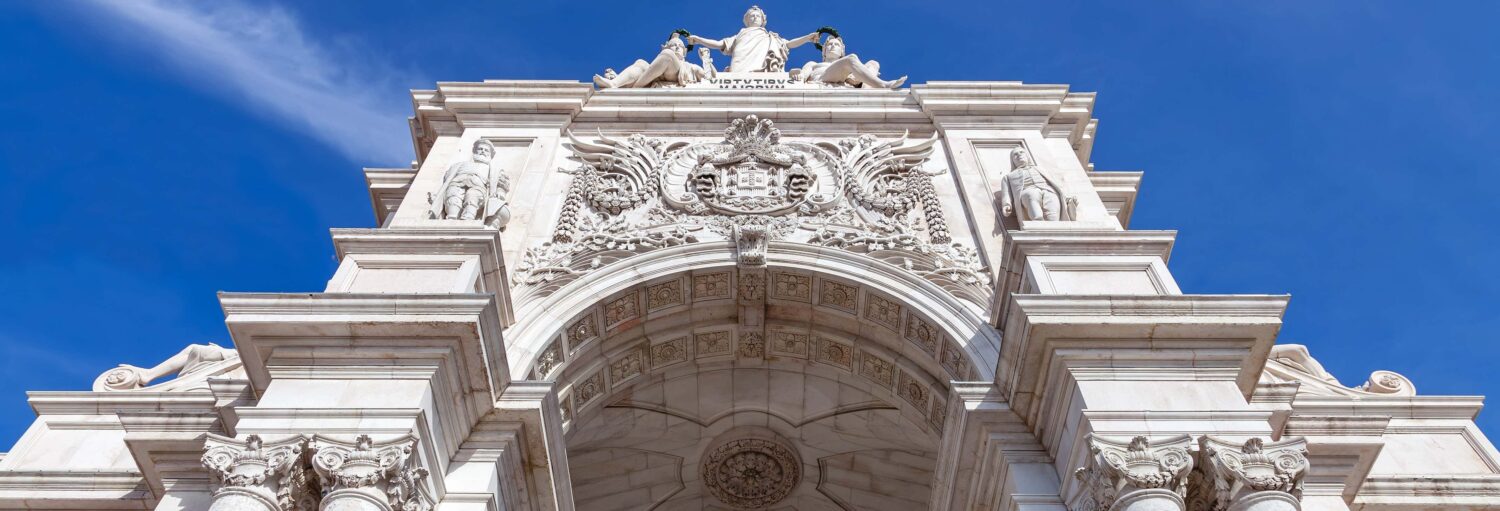 Arco triunfal de pedra branca com esculturas e entalhes decorativos, contrastando com um céu azul claro. Imagem para ilustrar o post sobre ingresso do Arco da Rua Augusta.