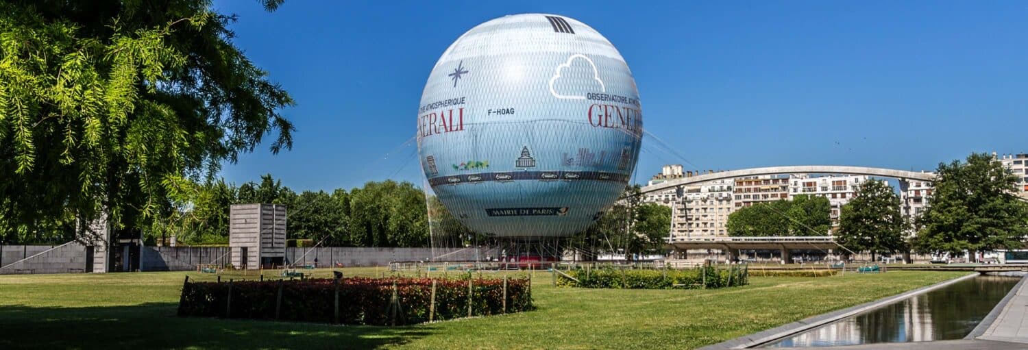Grande balão de ar quente preso com anúncios flutuando sobre uma área gramada de parque, com prédios ao fundo, ilustrando a página de ingresso do Ballon de Paris Generali