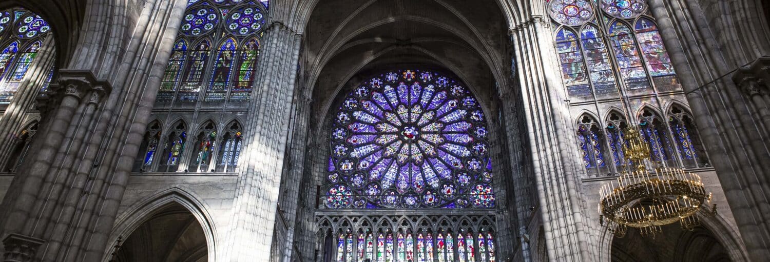 Vitral com rosáceas e arcos dentro de uma catedral gótica, com luz entrando e um lustre à direita. A imagem ilustra o post sobre ingresso da Basílica de Saint-Denis.