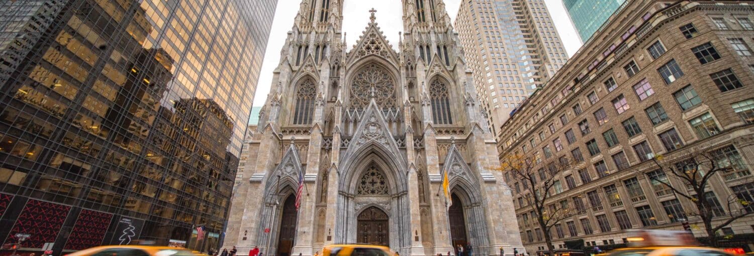 Fachada da Catedral de São Patrício, na cidade de Nova York, com prédios altos e táxis amarelos em primeiro plano. A foto é capa do post Ingresso da catedral de St. Patrick com audioguia.
