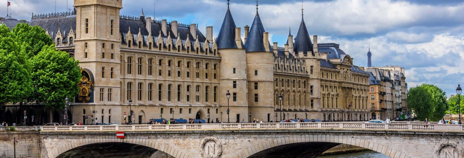 Uma ponte de pedra cruza o Rio Sena em frente ao histórico edifício Conciergerie, em Paris, sob um céu nublado. A imagem ilustra o post sobre ingresso da Conciergerie.