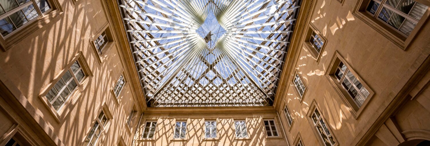 Teto de vidro com design geométrico projetando sombras em um pátio cercado por paredes e janelas de pedra bege para ilustrar o passeio de ingresso dos apartamentos do Intendente do Hôtel de la Marine