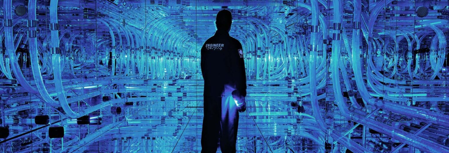 Uma pessoa vestindo um macacão de "Engenheiro" está em uma sala futurista, com iluminação azul, cheia de cabos e superfícies reflexivas. A foto é capa do post Ingresso do Mercer Labs.