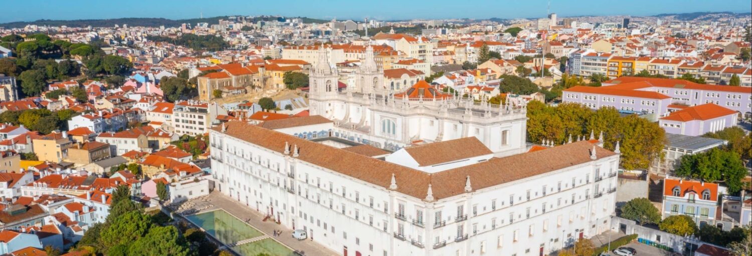 Vista aérea de um grande palácio histórico branco com telhado de telhas vermelhas, cercado por prédios urbanos e vegetação. Imagem para ilustrar post sobre ingresso do mosteiro de São Vicente de Fora.