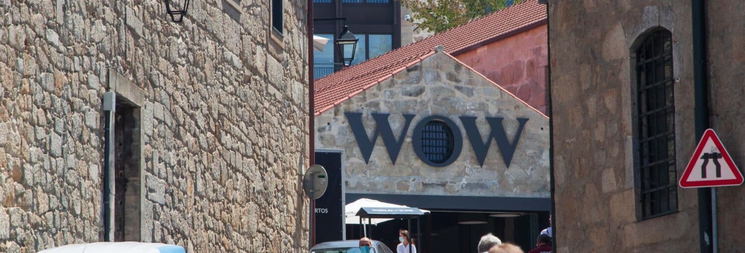 Prédios de pedra se alinham em uma rua estreita, com uma placa dizendo "WOW" em um prédio ao fundo. A foto é capa do post de Ingresso de um museu do WOW Porto.