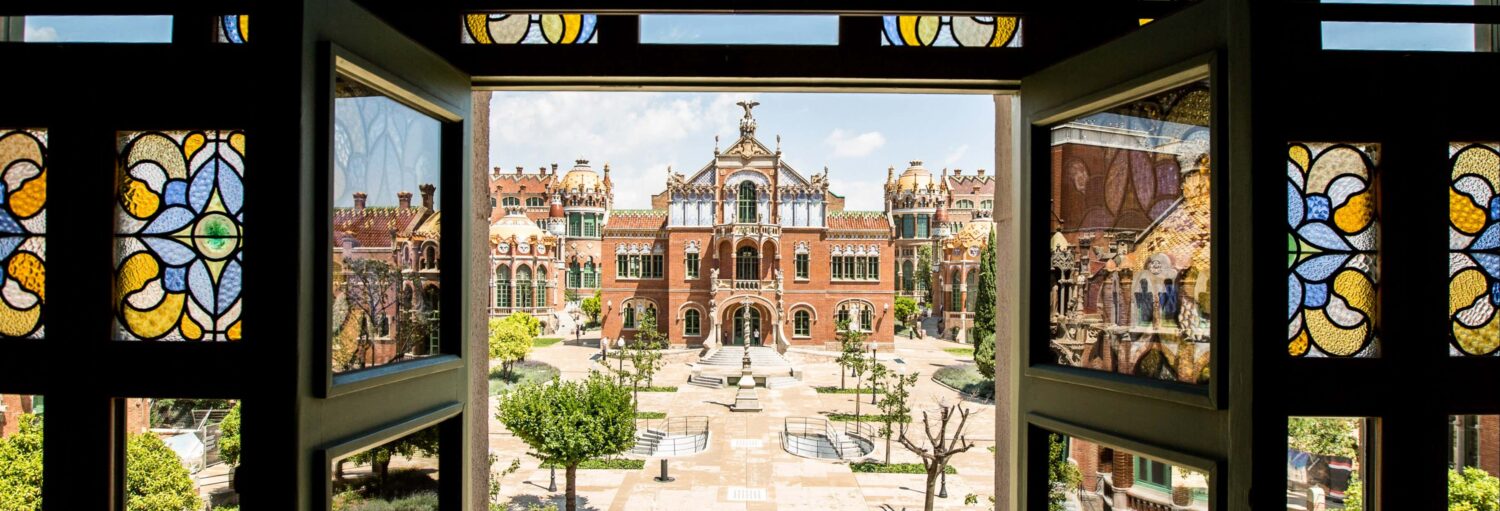 Vista de um pátio histórico e de um edifício ornamentado de tijolos vermelhos, visto através de vitrais. A imagem ilustra o post sobre Ingresso do Recinte Modernista de Sant Pau.