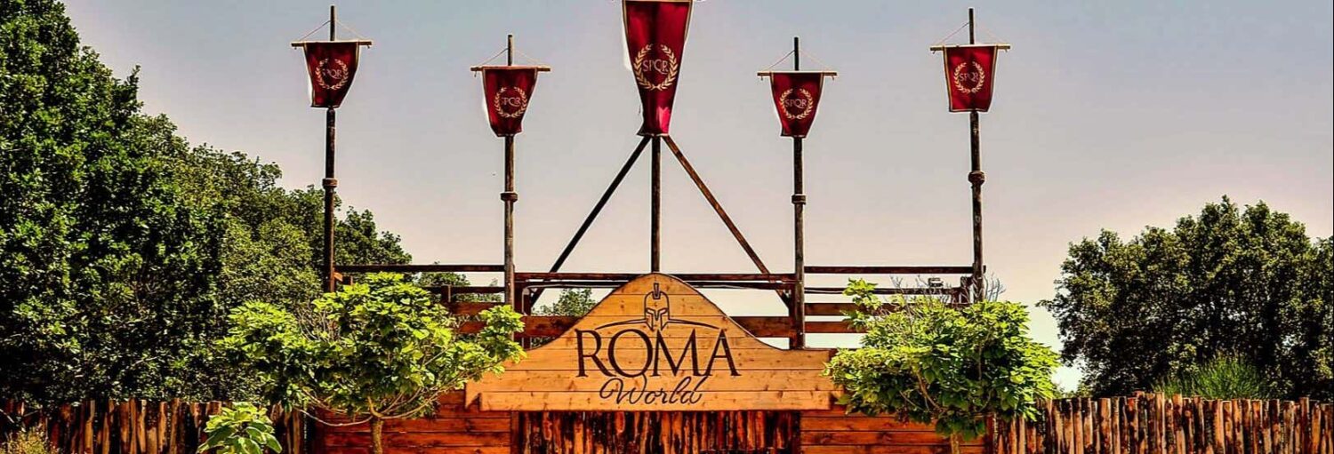 Entrada para o parque temático Roma World com cerca de madeira, faixas vermelhas, vegetação e a placa "ROMA World" no centro, que pode ser visitado com o Ingresso do Roma World