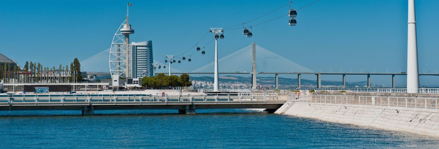 Um rio com um teleférico, uma ponte e edifícios modernos ao fundo, sob um céu azul claro. Imagem para ilustrar post sobre ingresso do teleférico de Lisboa.