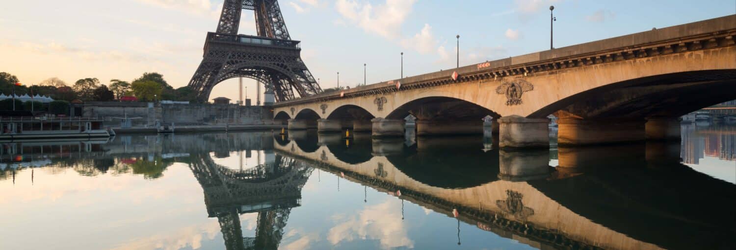 A Torre Eiffel e uma ponte de pedra refletem-se nas águas calmas do Rio Sena ao nascer do sol. A imagem ilustra o post sobre Ingresso da Torre Eiffel + Cruzeiro pelo Sena.